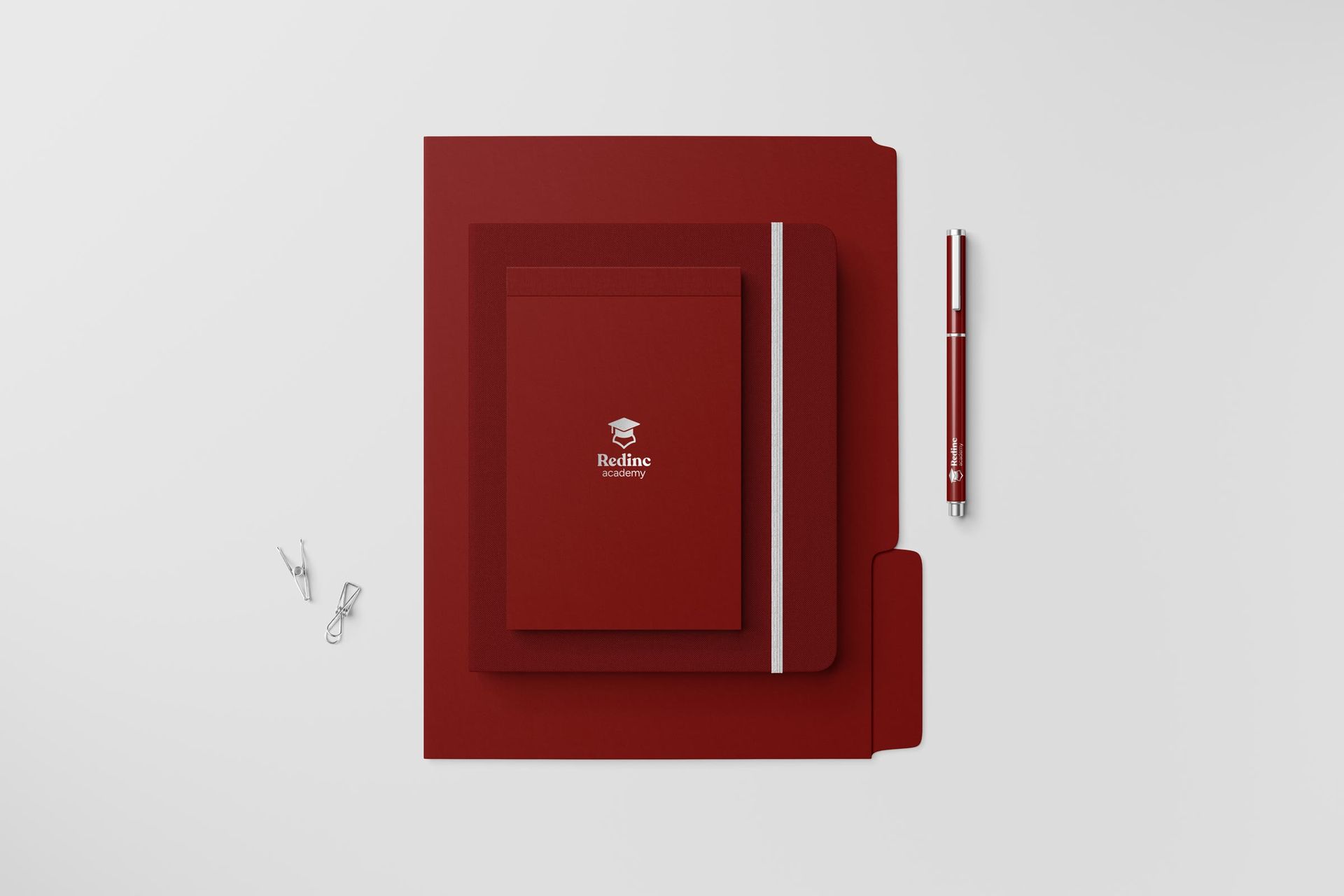 Mockup of the Redinc stationery set: a burgundy folder, notebook and notepad stacked flat on a pale grey surface, the white cap mark and Redinc academy wordmark centred on the notepad, a matching burgundy pen and two paper clips beside them