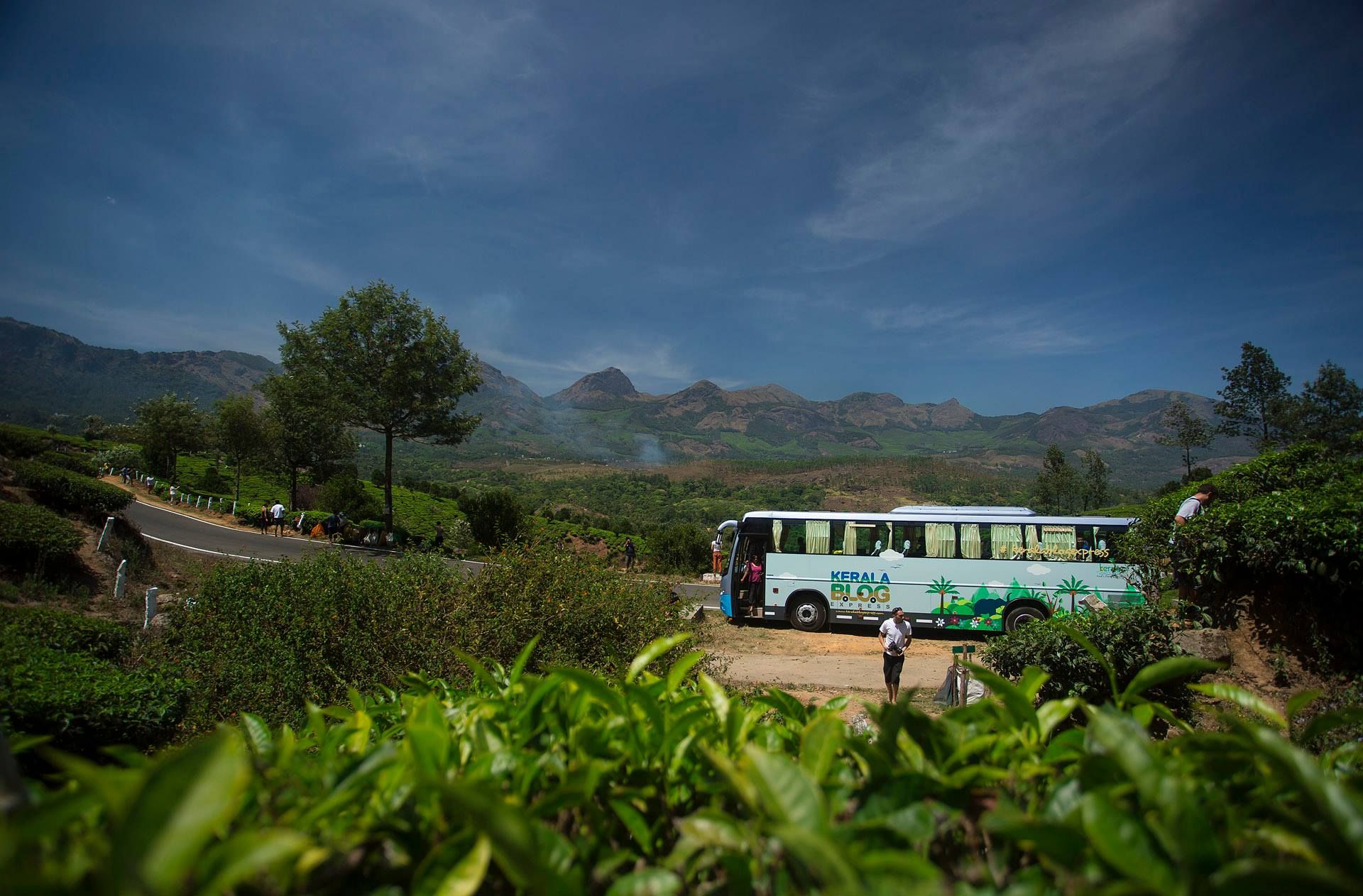 The Kerala Blog Express bus parked beside a hill road among tea plantations, with mountains behind
